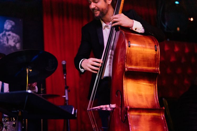 A white man wearing a white shirt and black blazer plays a double bass. He is standing on a stage.